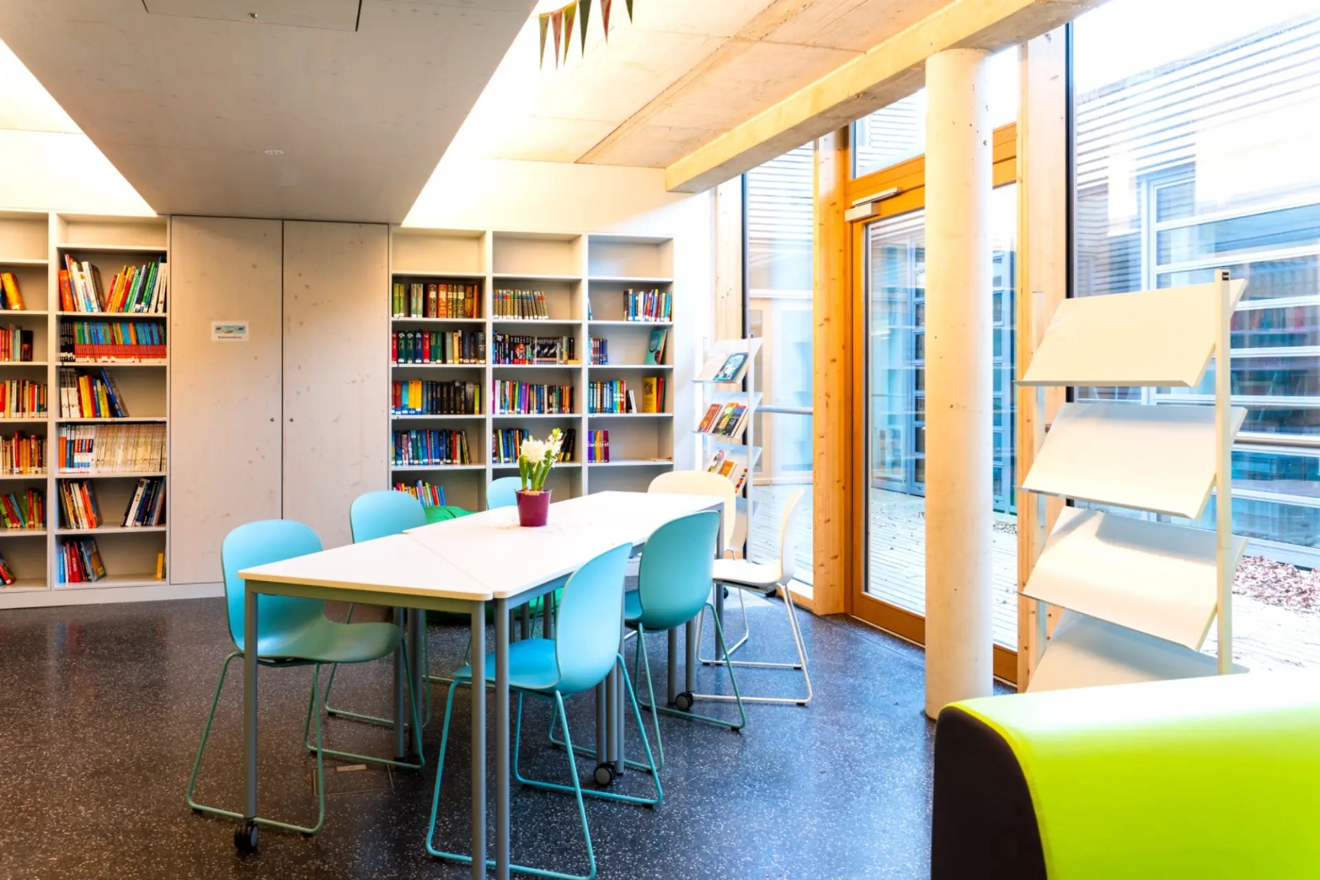 Ein moderner Schulhof mit einem zweistöckigen Schulgebäude im Hintergrund. Das Gebäude kombiniert graue Backsteinfassade mit horizontaler Holzverkleidung und großen Glasfronten. Im Vordergrund führt eine breite Betontreppe mit Metallgeländern auf eine gepflasterte Ebene.