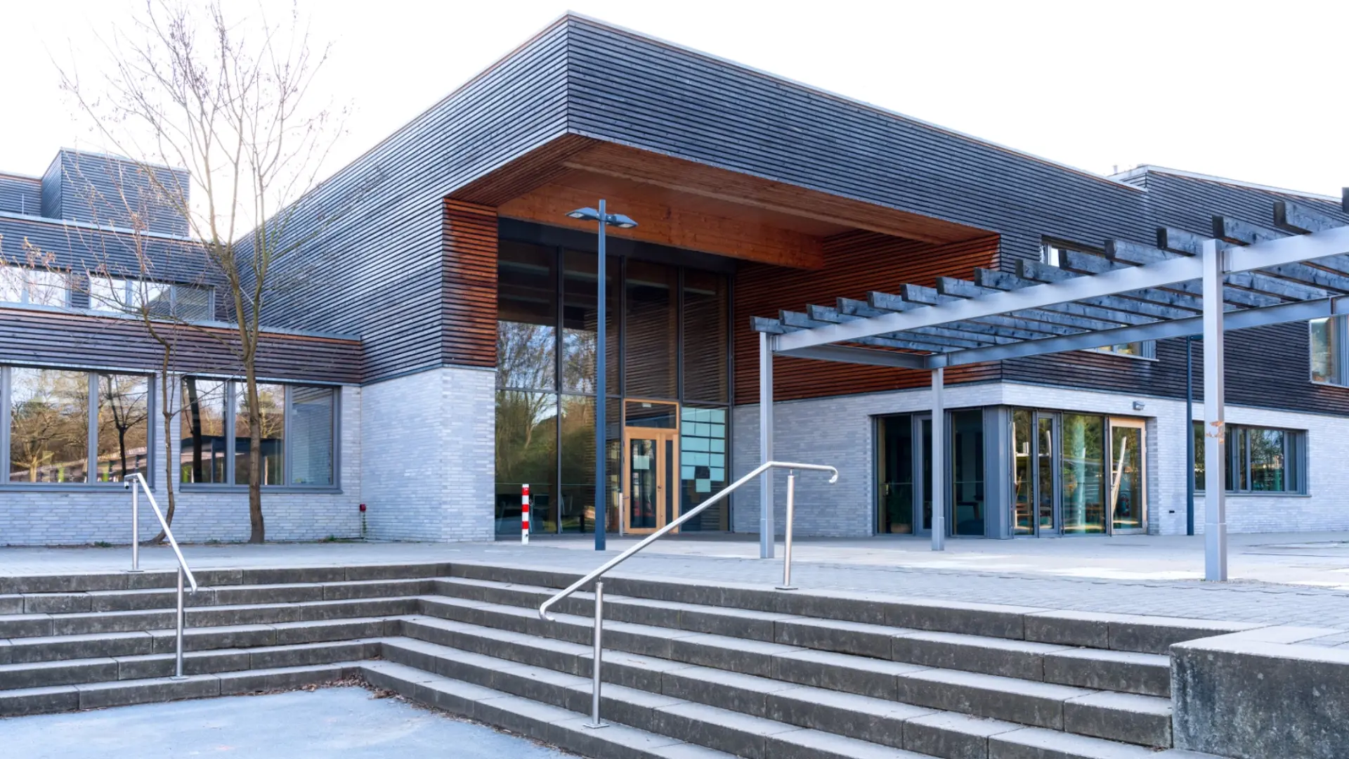 Ein moderner Schulhof mit einem zweistöckigen Schulgebäude im Hintergrund. Das Gebäude kombiniert graue Backsteinfassade mit horizontaler Holzverkleidung und großen Glasfronten. Im Vordergrund führt eine breite Betontreppe mit Metallgeländern auf eine gepflasterte Ebene.