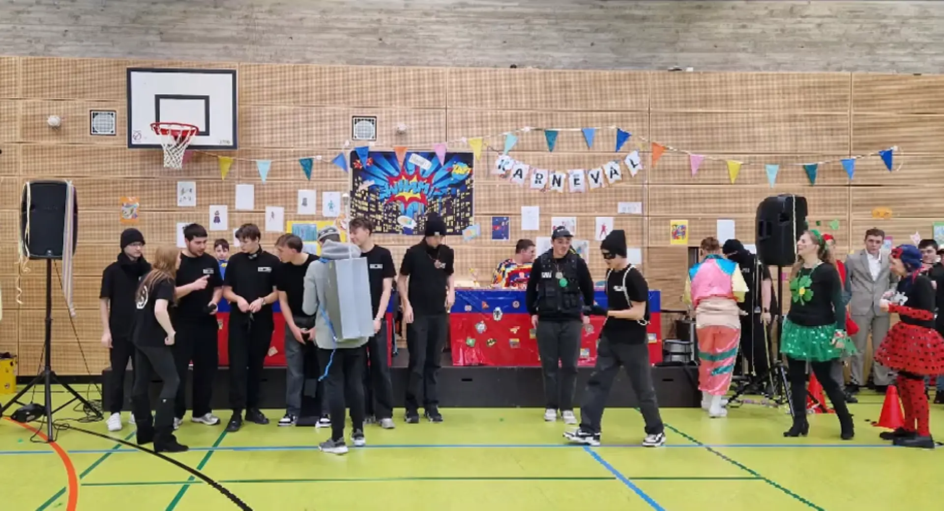 Mehrere gleich kostümierte Schüler*innen besprechen etwas bzgl. ihres Auftritts. Im Hintergrund sieht man bunte Girlande und Bilder an der Wand der Turnhalle, die Teil der Karnevalsdekoration sind. 