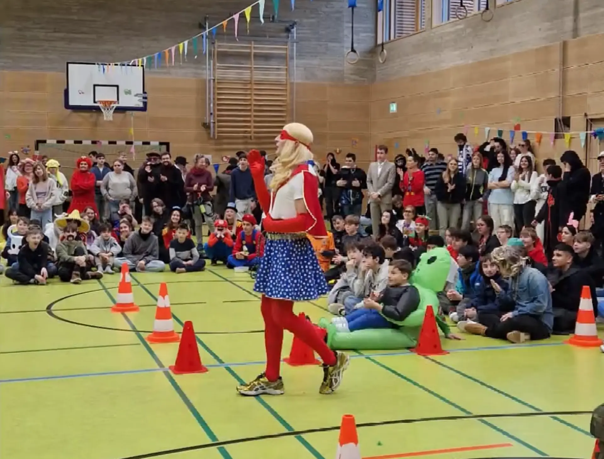 Eine Lehrkraft, die als Wonder Woman verkleidet ist, moderiert am Mikrofon in der dekorierten Turnhalle. Drum-herum stehen und sitzen viele Schüler*innen und Mitarbeitende und hören zu. 