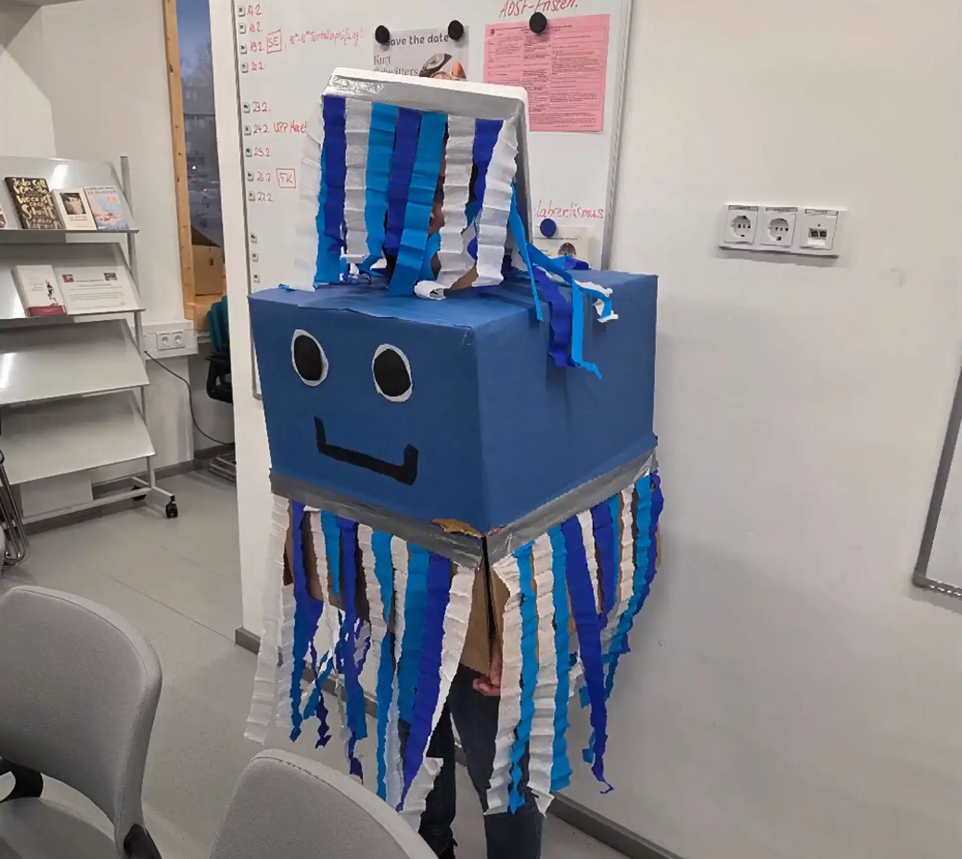 Ein kostümierter Mensch im Lehrer*innen-Zimmer. Das Kostüm besteht aus einem blauen Karton mit Augen und einem lächelnden Mund, nach unten hängen blauen und weiße Girlanden. 
