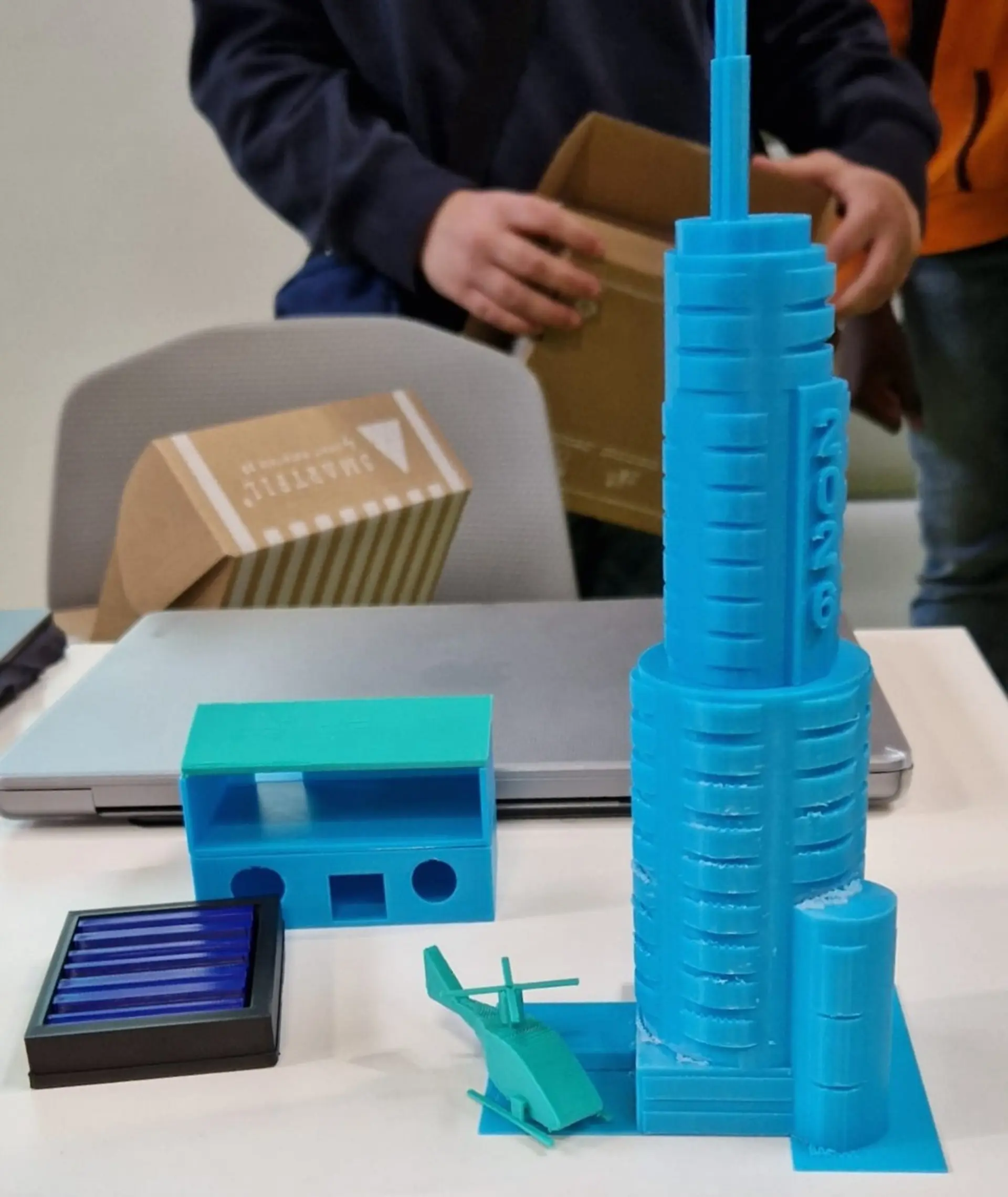 Mehrere 3D-gedruckte Kunststoffmodelle auf einem Tisch, darunter ein hoher Turm mit der Aufschrift 2026, ein kleiner Hubschrauber und geometrische Gebäudeformen während eines MINT-Workshops.