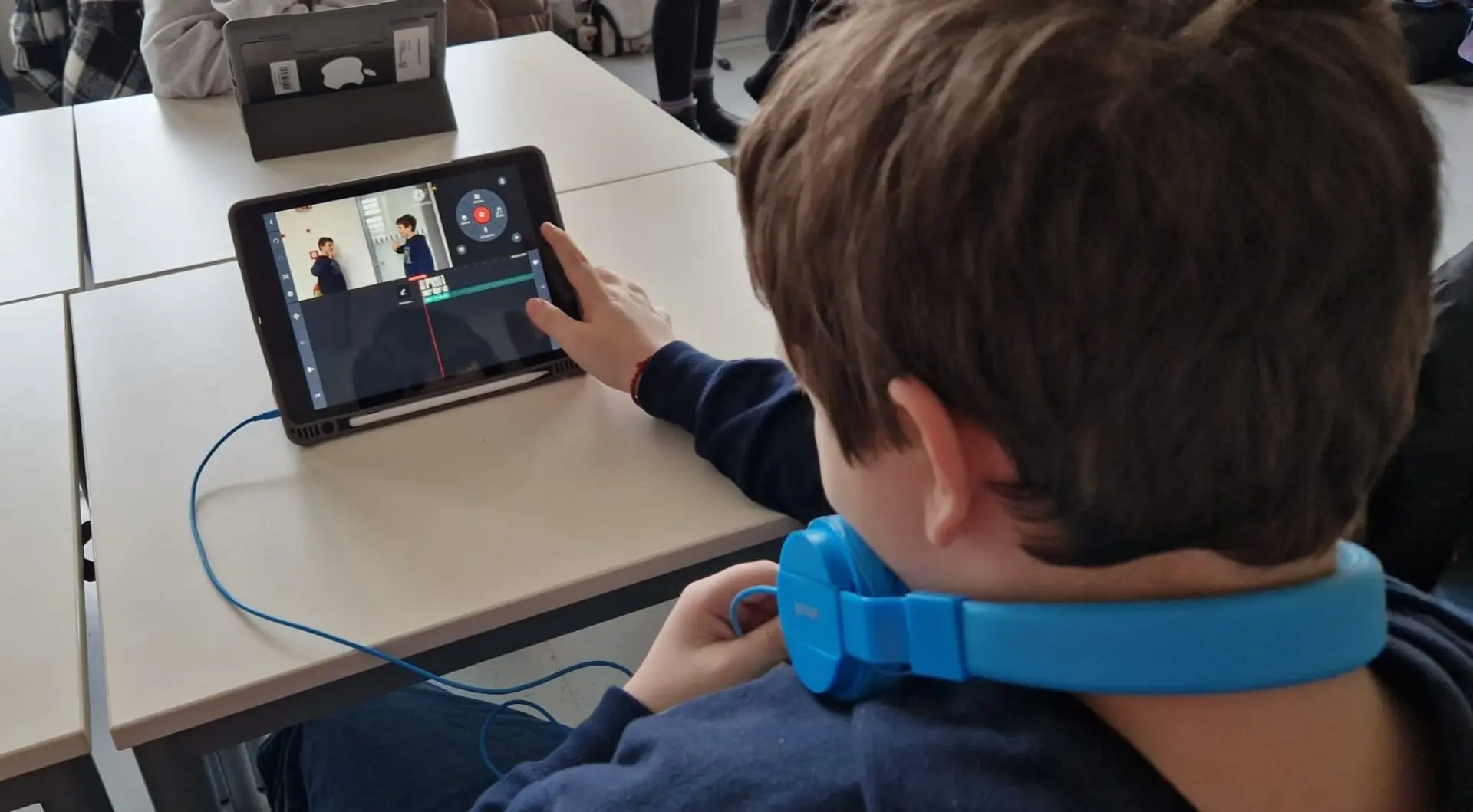 Rückansicht eines Schülers mit Kopfhörern um den Hals, der an einem Tablet ein Video schneidet. Auf dem Bildschirm ist eine Videobearbeitungs-Software mit einer Zeitleiste und einer Filmszene zu sehen, während eines MINT-Workshops zur Filmproduktion.