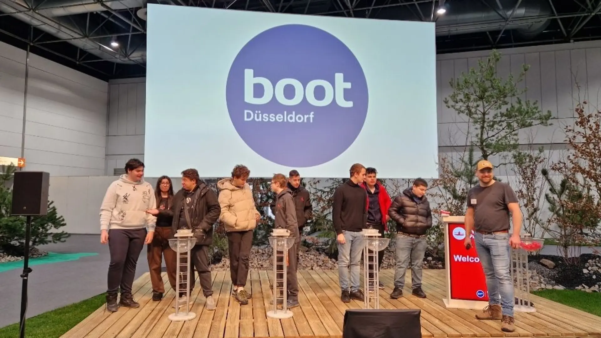 Mehrere Schüler*innen auf einer Bühne auf dem Messegelände. Sie sind in kleinen Grüppchen aufgeteilt und jede Gruppe bespricht etwas. An der Seite steht ein Erwachsener als Moderator, im Hintergrund sieht man ein großes Plakat mit der Schrift "boot Düsseldorf". 