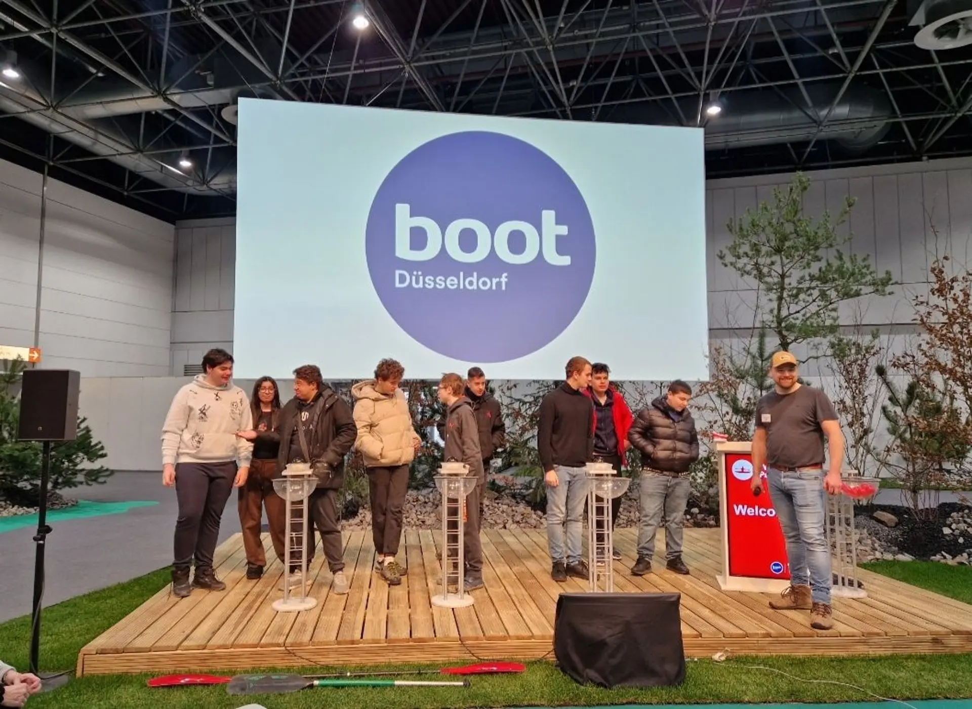 Mehrere Schüler*innen auf einer Bühne auf dem Messegelände. Sie sind in kleinen Grüppchen aufgeteilt und jede Gruppe bespricht etwas. An der Seite steht ein Erwachsener als Moderator, im Hintergrund sieht man ein großes Plakat mit der Schrift "boot Düsseldorf". 